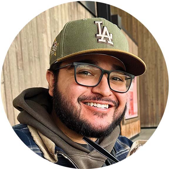 A smiling man with glasses, a beard, and a mustache wearing a green Los Angeles Dodgers baseball cap, a dark hoodie, and a jacket.