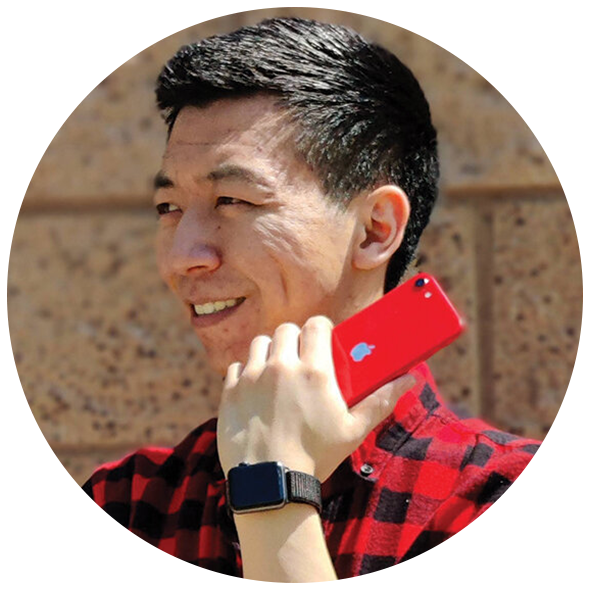 A man smiling and holding a red iPhone to his ear, wearing a red and black plaid shirt and a smartwatch, outdoors with a brick wall in the background.