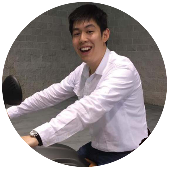 A young man with short black hair smiling while sitting on a scooter, wearing a white long-sleeve shirt, against a brick wall background.