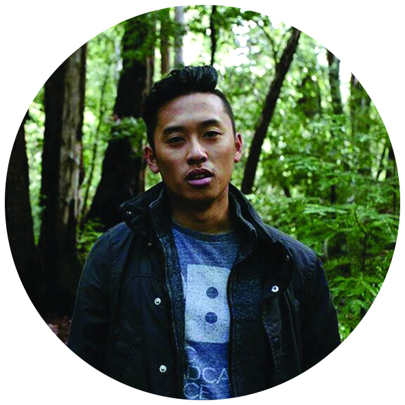 A young man with black hair wearing a black jacket and a blue graphic t-shirt standing in a forest with green trees and foliage in the background.