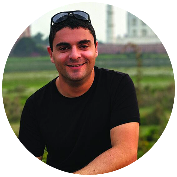 A smiling young man with short dark hair, wearing a black t-shirt and sunglasses on his head, outdoors with green fields and buildings in the background.